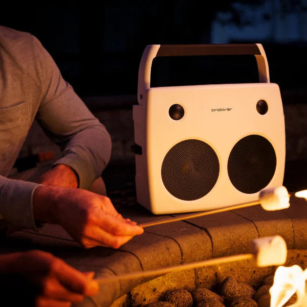 Portable white speaker on a stone surface with a person roasting marshmallows by a fire.